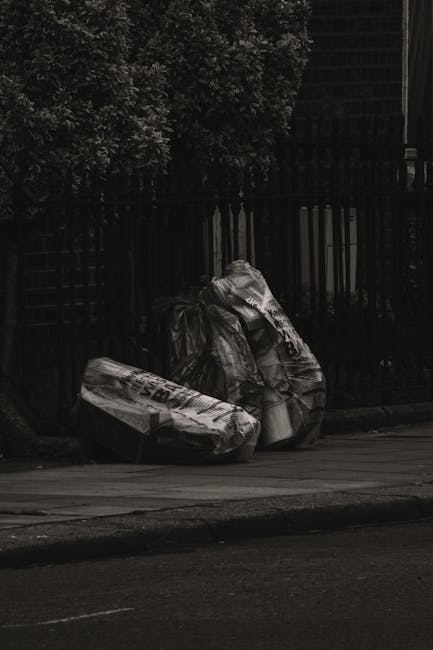 The image depicts two large black plastic garbage bags filled with waste, resting on a sidewalk curb in an urban residential area. One bag is partially leaning against the other, with crumpled and slightly torn plastic visible at the folds and edges. The bags are placed near a black metal fence with vertical slats, which runs along the background behind dense, leafy foliage. The scene appears to be at dusk or nighttime, with dim lighting creating subtle highlights on the surface of the bags and the pavement. The setting suggests a typical street scene where residents or private waste disposal services, such as Rubbish Clearance Edgware, might handle rubbish collection outside of council-managed services, emphasizing the importance of professional rubbish removal including on-site clearance and independent waste handling options. The overall atmosphere is calm and orderly, although the image captures waste awaiting collection, highlighting the relevance of reliable rubbish removal services in maintaining urban cleanliness and convenience.