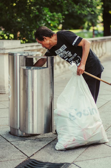 A middle-aged man with dark, slightly wavy hair is seen wearing a black T-shirt with white text and dark pants. He is bending over to dispose of rubbish into a cylindrical, stainless steel outdoor public trash bin with a rounded top and a rectangular opening at the front. The man is holding a large white plastic bag filled with waste in his left hand, while his right hand is reaching into the bin to deposit trash. The scene is set on a paved walkway, with a stone balustrade running along the edge in the background, surrounded by lush green trees and foliage. The lighting suggests daytime, with natural sunlight illuminating the scene. The environment appears clean and well-maintained, and this image aligns with concepts of private waste disposal and alternative rubbish collection methods, as handled by waste management services like Rubbish Clearance Edgware.