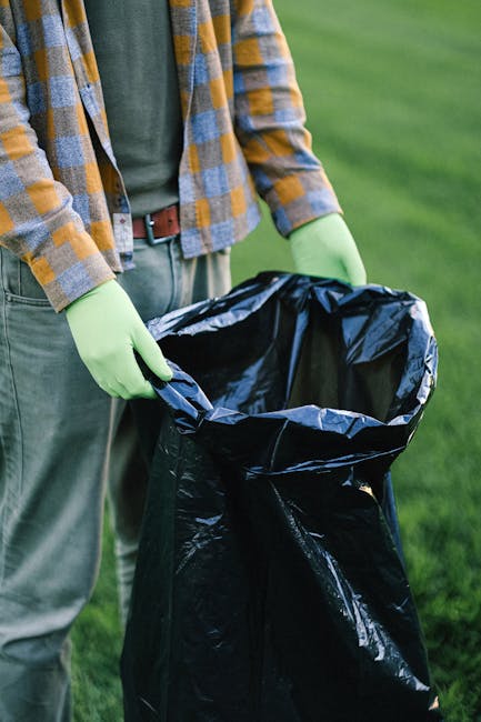 The image depicts two large black plastic garbage bags filled with waste, resting on a sidewalk curb in an urban residential area. One bag is partially leaning against the other, with crumpled and slightly torn plastic visible at the folds and edges. The bags are placed near a black metal fence with vertical slats, which runs along the background behind dense, leafy foliage. The scene appears to be at dusk or nighttime, with dim lighting creating subtle highlights on the surface of the bags and the pavement. The setting suggests a typical street scene where residents or private waste disposal services, such as Rubbish Clearance Edgware, might handle rubbish collection outside of council-managed services, emphasizing the importance of professional rubbish removal including on-site clearance and independent waste handling options. The overall atmosphere is calm and orderly, although the image captures waste awaiting collection, highlighting the relevance of reliable rubbish removal services in maintaining urban cleanliness and convenience.