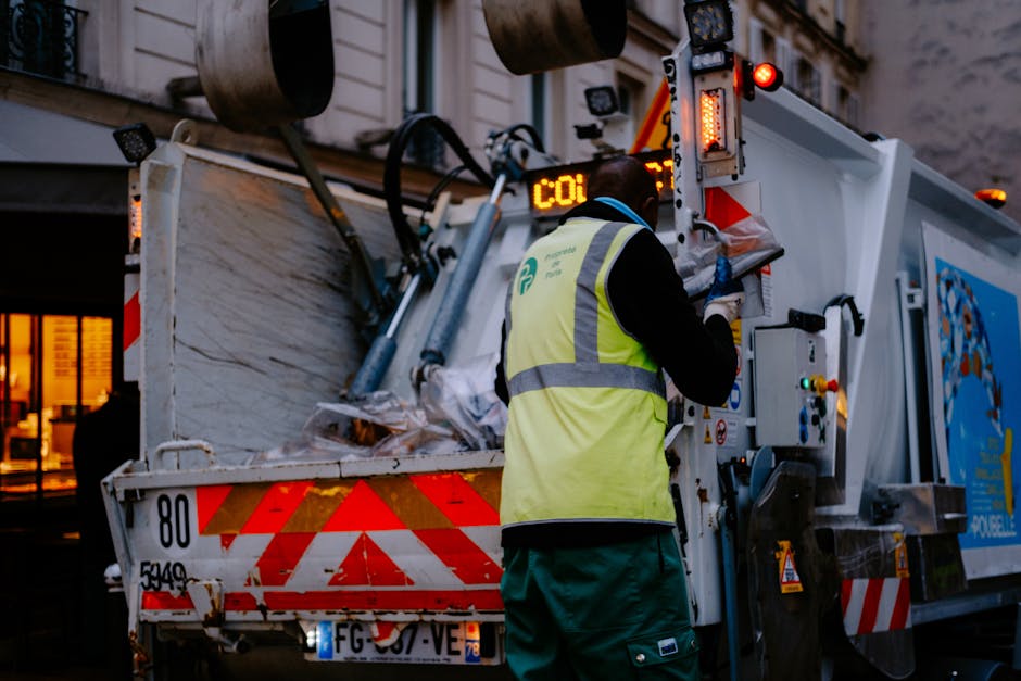 A worker wearing a high-visibility yellow vest with reflective stripes and green work trousers is operating control panels on a large commercial waste collection vehicle parked on an urban street during dusk or evening. The vehicle's rear compartment is open, revealing a load of crumpled newspapers, paper waste, and other mixed recyclables stacked inside with visible textures of paper and cardboard. The vehicle has red and white chevron markings on its lower rear section, and its lighting is illuminated, casting a glow on the surrounding area. In the background, there are blurred buildings with lit windows, indicating a city environment. The worker appears to be engaged in managing or monitoring a rubbish collection or private waste disposal process, aligning with independent rubbish removal services often used for on-site clearance or alternative waste handling outside local authority collections. The scene emphasizes a professional waste management operation tailored for detailed rubbish removal tasks in Edgware or similar urban settings, with the company Rubbish Clearance Edgware providing tailored services for such waste handling.