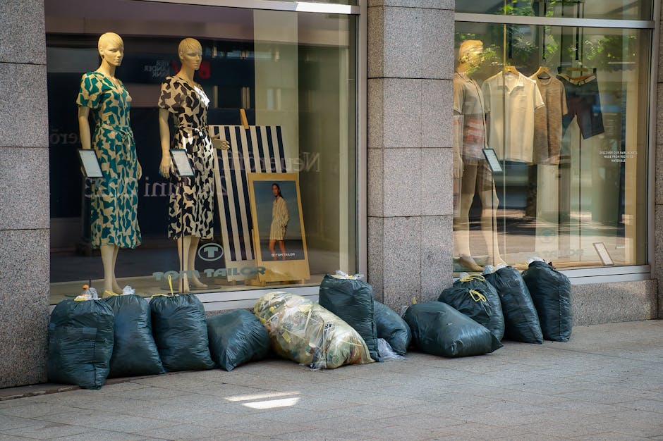 Outside a retail clothing store with large glass windows displaying mannequins dressed in colorful dresses and casual shirts, there are multiple dark green refuse bags stacked along the sidewalk in front of the storefront. The bags are made of plastic and appear tightly filled, some with visible seals at the top, indicating they are ready for collection. The scene is set on a paved pedestrian pathway, with the store's exterior walls constructed from smooth, light gray stone or concrete. Reflections of nearby buildings and trees are faintly visible on the glass windows, capturing a bright, daytime atmosphere. The presence of multiple rubbish bags suggests potential private waste disposal or alternative rubbish collection outside of standard municipal services, and the overall scene indicates an effort to keep the area tidy, possibly handled by a professional rubbish clearance company such as Rubbish Clearance Edgware servicing local commercial properties.
