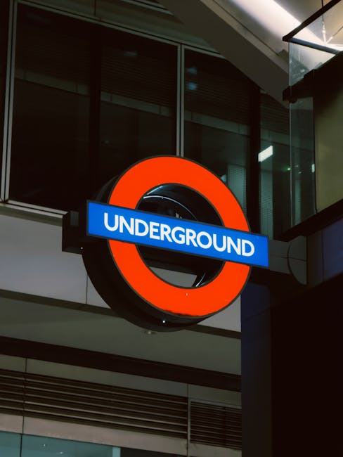 A circular London Underground sign featuring a bright orange outer ring and a central black circle with a horizontal blue bar across the middle displaying the word 'UNDERGROUND' in white text. The sign is mounted on a dark-colored metal bracket attached to a modern building with a glass façade. Behind the sign, there are large glass windows reflecting interior lighting and structural elements of the building. The scene suggests an urban environment near a public transport station, with the sign visible from street level or an entrance area, highlighting the familiar symbol of London's underground transit system. The lighting appears to be from ambient interior lights, casting subtle reflections on the glass surfaces.