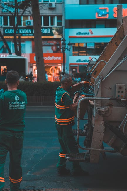 Two waste management workers operating a large urban rubbish collection vehicle on a city street during daylight hours. One worker, facing away, wears a dark green uniform with the logo and text 'Cankaya Belediyesi' on the back, indicating municipal waste services. The second worker, positioned beside the vehicle's hopper, is dressed in a high-visibility vest with orange reflective stripes and dark trousers, and appears to be engaging with controls or preparing to load or unload waste. The background shows a busy street scene with multi-storey retail buildings featuring logos and advertisements, including a Vodafone store and a Bankara branch, with trees lining the sidewalk. The scene captures a routine operation related to private or municipal rubbish collection on an urban thoroughfare, highlighting on-site waste handling as part of regular service operations.