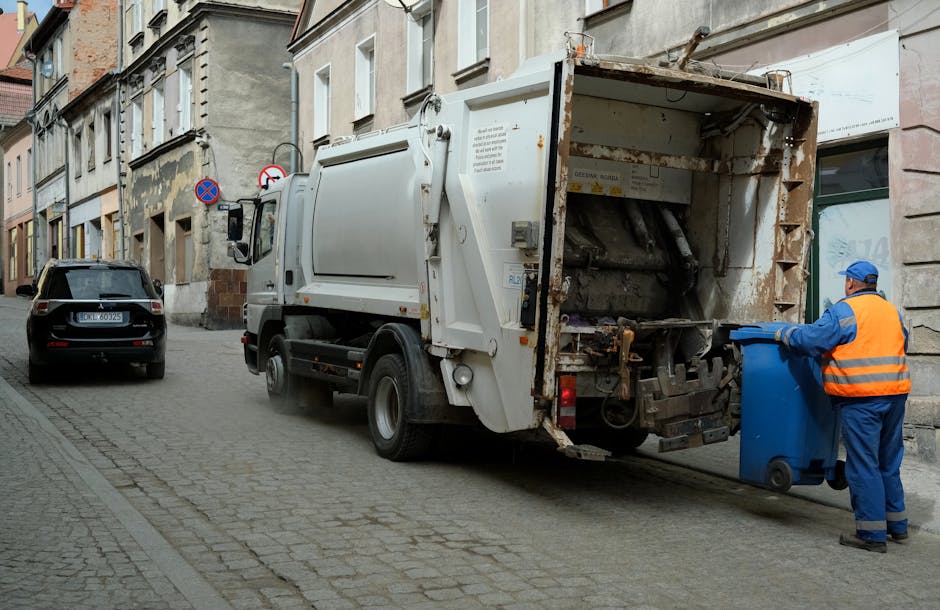 A street scene shows a white waste collection truck positioned on a cobblestone road beside a row of older residential buildings. The rear of the vehicle is open, revealing the mechanical compaction mechanism inside, which appears rusted and used. A worker dressed in blue overalls and an orange high-visibility vest, wearing a blue cap, is standing to the right of the truck, pushing a blue wheeled bin towards the open rear hatch. To the left of the truck, a black SUV is parked facing away from the camera, with the vehicle's number plate visible. The buildings in the background have a worn appearance with weathered facades and patchy paintwork, typical of an urban environment. The scene is lit by natural daylight, and the overall setting suggests an operational waste collection activity, potentially as part of an independent rubbish removal service like Rubbish Clearance Edgware, offering private waste handling or on-site clearance. Visible signs include a no parking restriction and a no entry sign mounted on lampposts along the street.