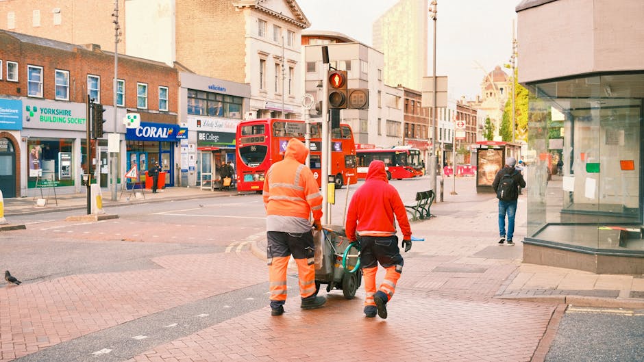 Two waste management workers operating a large urban rubbish collection vehicle on a city street during daylight hours. One worker, facing away, wears a dark green uniform with the logo and text 'Cankaya Belediyesi' on the back, indicating municipal waste services. The second worker, positioned beside the vehicle's hopper, is dressed in a high-visibility vest with orange reflective stripes and dark trousers, and appears to be engaging with controls or preparing to load or unload waste. The background shows a busy street scene with multi-storey retail buildings featuring logos and advertisements, including a Vodafone store and a Bankara branch, with trees lining the sidewalk. The scene captures a routine operation related to private or municipal rubbish collection on an urban thoroughfare, highlighting on-site waste handling as part of regular service operations.